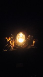 An altar lit by a bright lantern-candle in the dark, outside, with apples and drinking cup barely visible.