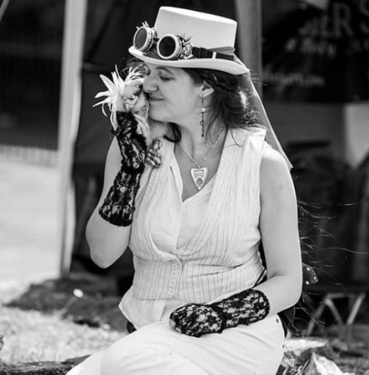black and white picture of a femme person in white, with white top hat, sat on a wall with a mini gryphon snuggled on their shoulder.