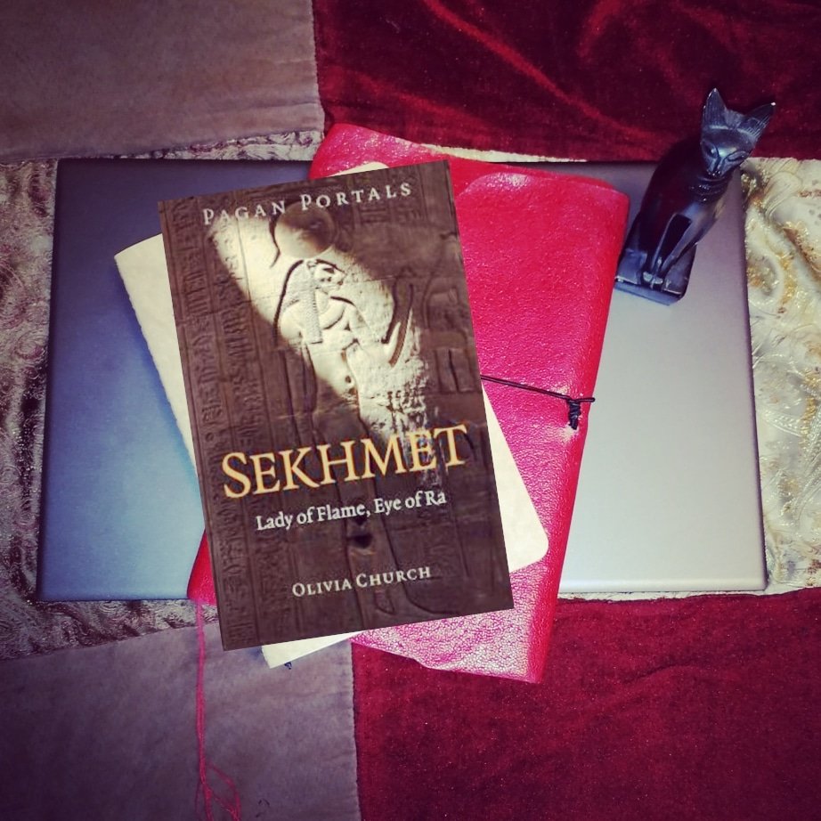 A pile of books on top of a laptop, "Pagan Portals: Sekhmet" on top, with a small black cat statuette, and a red and gold background.