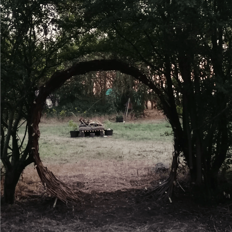 A circular doorway of willow between two hawthorn trees, framing a stone altar.